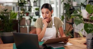 woman in a shop holding a clipboard looking at laptop